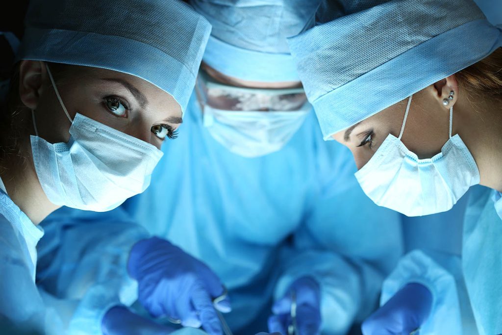 Three surgeons in blue gowns and masks are looking at the camera.